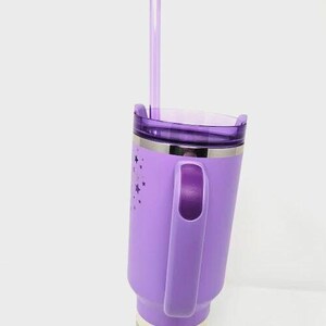 May include: A purple insulated tumbler with a handle and a clear straw. The tumbler has a silver band and a lid. The side of the tumbler is decorated with star shapes. The tumbler is designed to hold liquids.