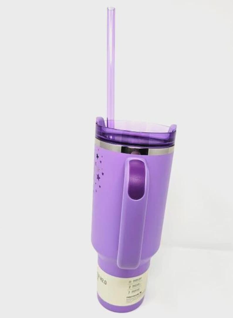 May include: A purple insulated tumbler with a handle and a clear straw. The tumbler has a silver band and a lid. The side of the tumbler is decorated with star shapes. The tumbler is designed to hold liquids.
