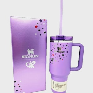 May include: A purple Stanley Quencher H2.0 tumbler with a handle and straw, next to a matching box. The tumbler and box feature a star pattern and the Stanley logo. The tumbler has a capacity of 40 oz.