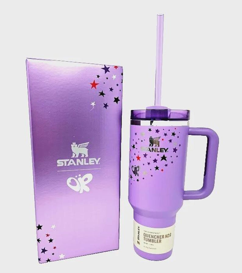 May include: A purple Stanley Quencher H2.0 tumbler with a handle and straw, next to a matching box. The tumbler and box feature a star pattern and the Stanley logo. The tumbler has a capacity of 40 oz.