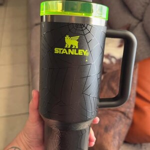 Op de afbeelding: Zwarte Stanley-beker met een spinnenwebontwerp, een neon groen deksel en een rietje. De beker heeft een grote handgreep en het Stanley-logo in neon groen. Deze ge&iuml;soleerde beker is ontworpen om dranken koud of warm te houden.