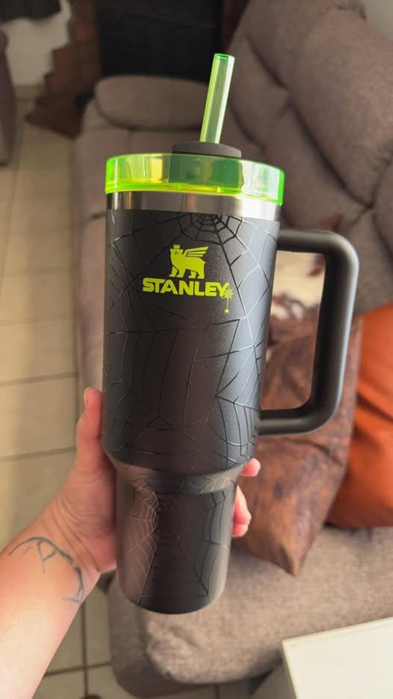 Op de afbeelding: Zwarte Stanley-beker met een spinnenwebontwerp, een neon groen deksel en een rietje. De beker heeft een grote handgreep en het Stanley-logo in neon groen. Deze ge&iuml;soleerde beker is ontworpen om dranken koud of warm te houden.