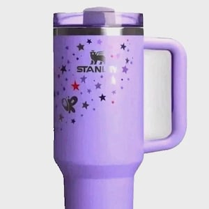 May include: A lavender Stanley tumbler with a handle and a lid. The tumbler is decorated with a pattern of stars and butterflies. The Stanley logo is visible on the side.