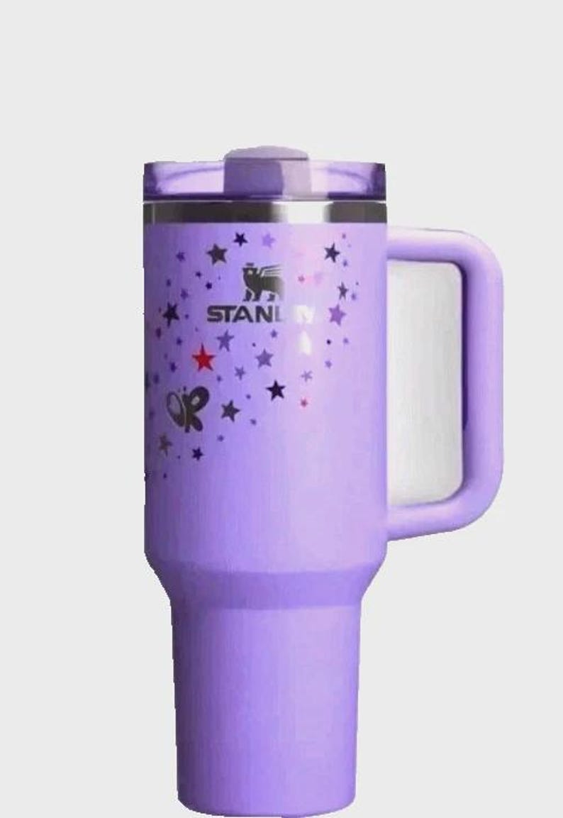 May include: A lavender Stanley tumbler with a handle and a lid. The tumbler is decorated with a pattern of stars and butterflies. The Stanley logo is visible on the side.