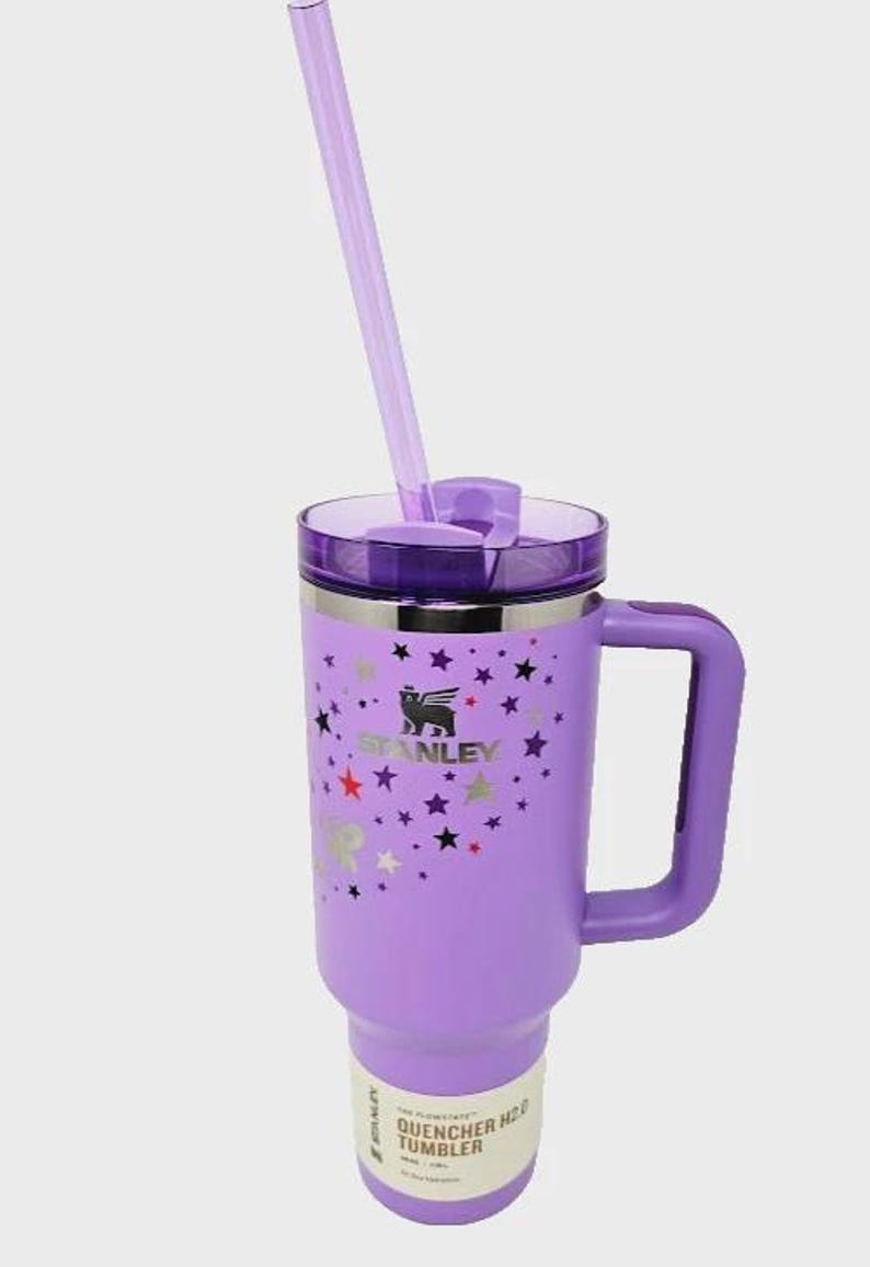 May include: A purple Stanley Quencher H2.0 tumbler with a handle and a clear straw. The tumbler features a lid, a handle, and a design of stars and the Stanley logo. The tumbler is designed for cold beverages.
