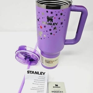 May include: A purple Stanley Quencher H2.0 tumbler with a handle, lid, and straw. The tumbler has a silver rim and is decorated with star graphics. Includes care instructions and a product information card.