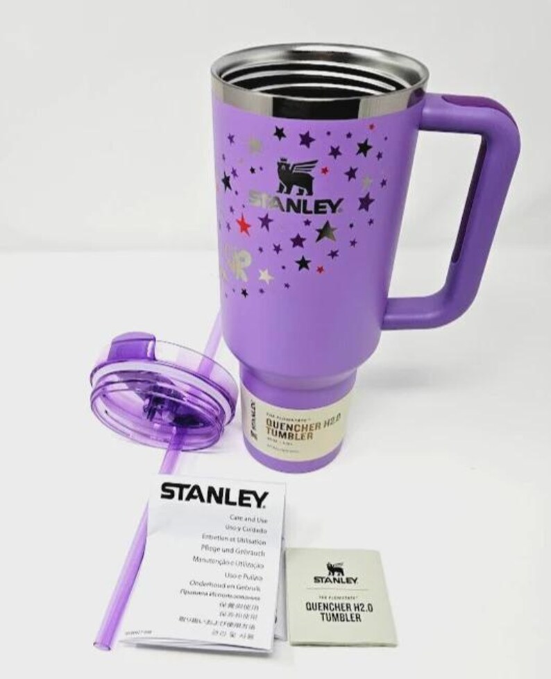 May include: A purple Stanley Quencher H2.0 tumbler with a handle, lid, and straw. The tumbler has a silver rim and is decorated with star graphics. Includes care instructions and a product information card.