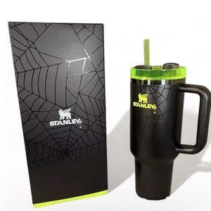 Op de afbeelding: Zwarte Stanley-beker met een handvat en een groen deksel, naast een bijpassende zwarte doos. Zowel de beker als de doos hebben een spinnenwebontwerp. Het Stanley-logo is op beide items zichtbaar.
