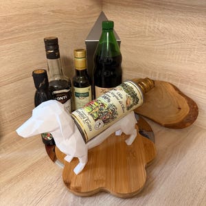 Puede incluir: Una colecci&oacute;n de botellas de aceite de oliva y vinagre, incluyendo una botella verde y una lata de aceite de oliva, exhibidas con un perro salchicha de papel blanco sobre una tabla de cortar de madera. La lata de aceite de oliva dice "Olio Extra Vergine di Oliva".