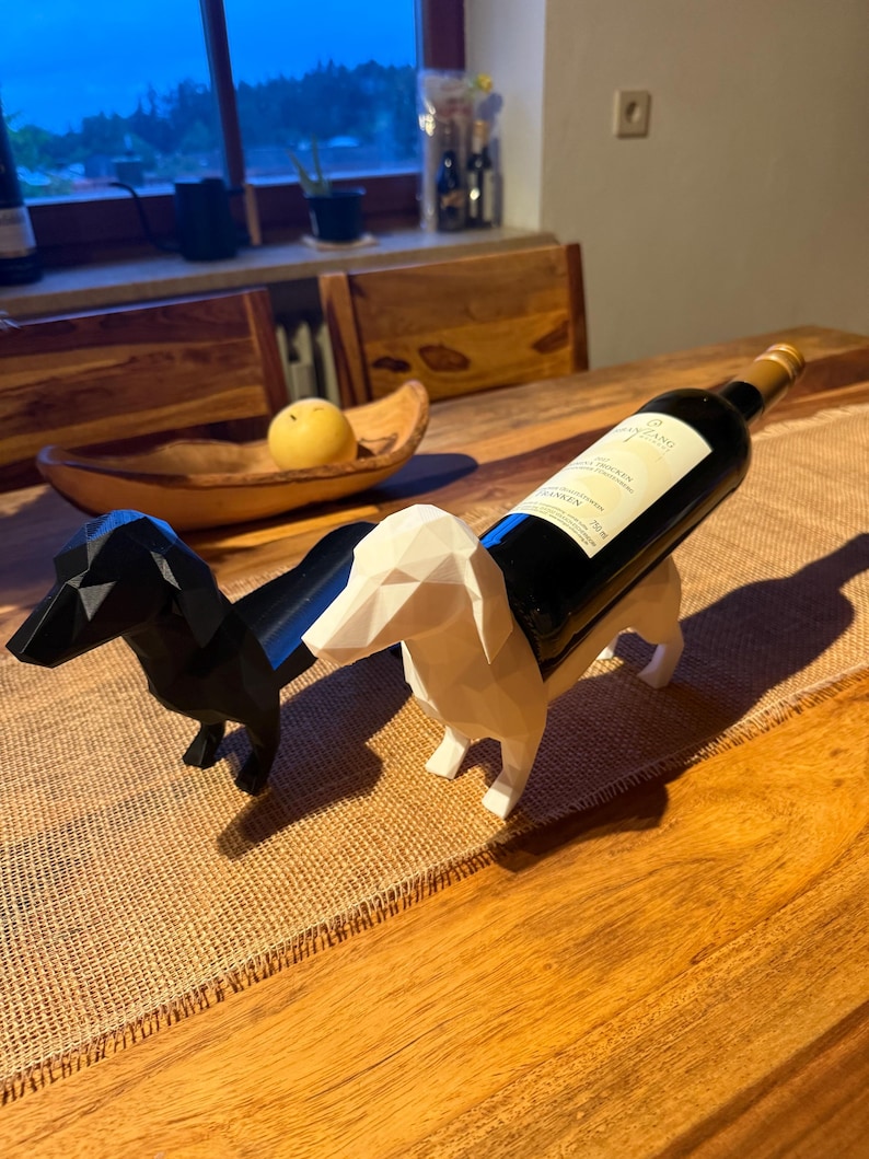 Puede incluir: Un soporte para botellas de vino con forma de perro salchicha en blanco y negro. Los perros salchicha est&aacute;n hechos de formas geom&eacute;tricas y sostienen una botella de vino. La botella es oscura con un cuello dorado y una etiqueta blanca. El soporte est&aacute; sobre una mesa de madera.