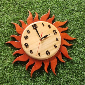 May include: A wooden clock shaped like a sun with orange rays. The clock face is light wood with black numbers and hands.