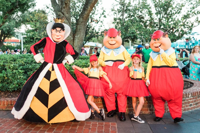 May include: The Queen of Hearts, the Mad Hatter, and the March Hare from Alice in Wonderland stand with two children dressed as Alice. The Queen is wearing a red and black dress with a white collar and a gold crown. The Mad Hatter and the March Hare are wearing red and yellow costumes with red hats. The children are wearing red and yellow dresses with red hats.