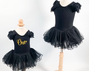 May include: Two black tulle tutus with black leotards on display mannequins. The smaller tutu has gold glitter lettering that says "One".