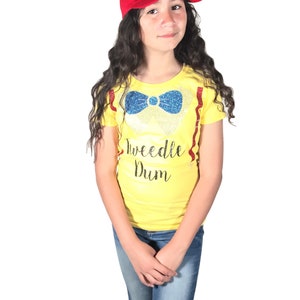 May include: A young person wearing a yellow t-shirt with a blue bow tie and the text "Tweedle Dum" in black. They are also wearing a red baseball cap with a yellow propeller on top and blue jeans.