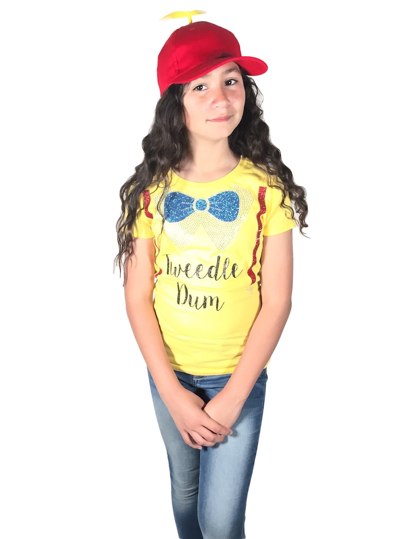 May include: A young person wearing a yellow t-shirt with a blue bow tie and the text "Tweedle Dum" in black. They are also wearing a red baseball cap with a yellow propeller on top and blue jeans.