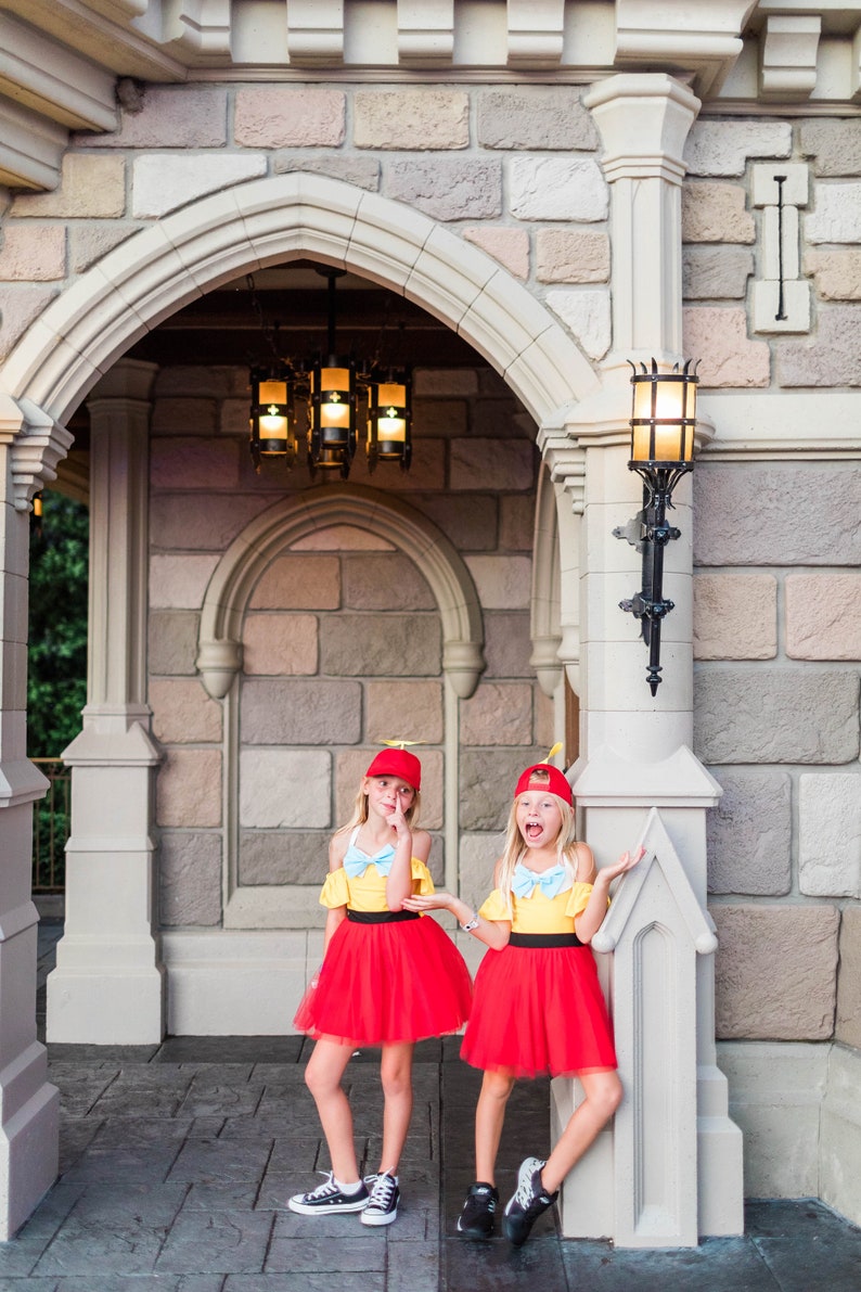 May include: Two girls dressed as Disney's  'Toy Story' characters, Woody and Jessie, stand in front of a stone archway. They are wearing red tulle skirts, yellow tops, and red hats with yellow accents. The girls are smiling and posing for a photo.