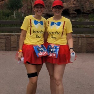May include: Two women wearing yellow shirts with the text "Tweedle Dee" and "Tweedle Dum" on them. They are wearing red tulle skirts and red hats with propellers. They are both wearing running shoes and have race bibs on.