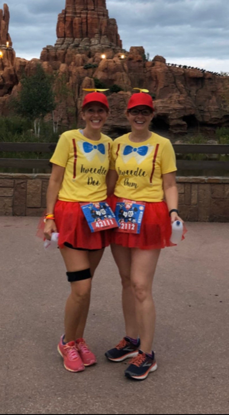 May include: Two women wearing yellow shirts with the text "Tweedle Dee" and "Tweedle Dum" on them. They are wearing red tulle skirts and red hats with propellers. They are both wearing running shoes and have race bibs on.