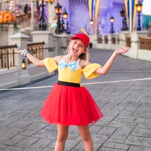 May include: A young girl dressed as a character from the Disney movie Pinocchio. She is wearing a yellow and red dress with a blue bow, a red hat with a propeller, and black sneakers. She is standing in front of Cinderella's Castle at Walt Disney World.