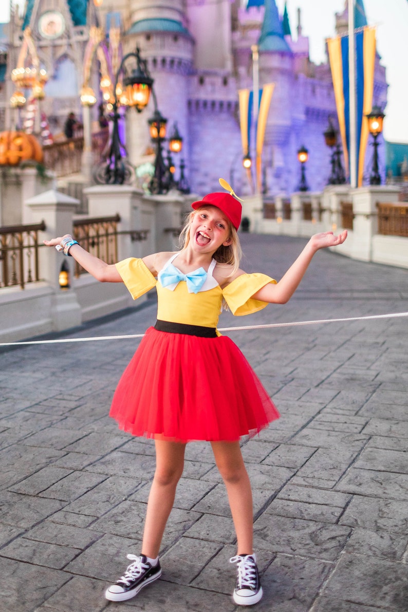 May include: A young girl dressed as a character from the Disney movie Pinocchio. She is wearing a yellow and red dress with a blue bow, a red hat with a propeller, and black sneakers. She is standing in front of Cinderella's Castle at Walt Disney World.