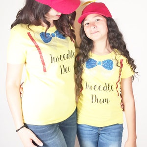 May include: Two people wearing yellow t-shirts with the text "Tweedle Dee" and "Tweedle Dum" on them. They are both wearing red baseball caps with yellow propeller accents.