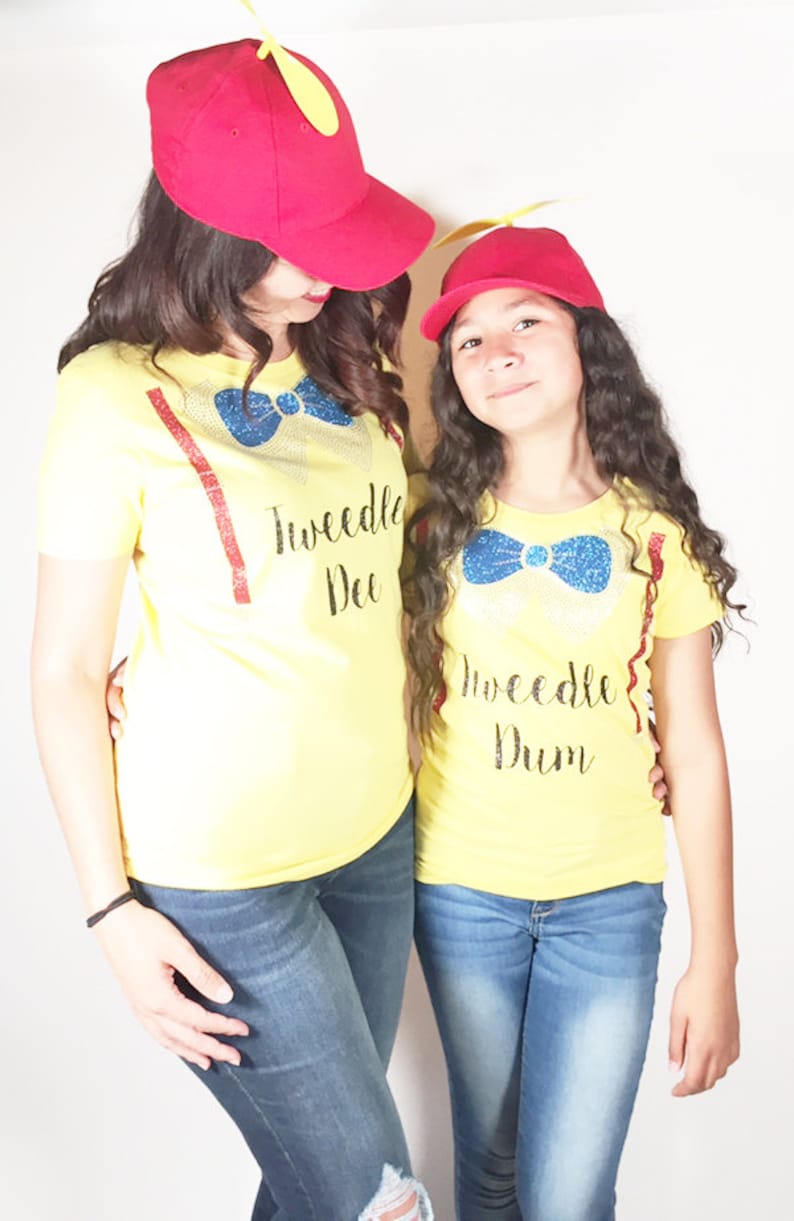 May include: Two people wearing yellow t-shirts with the text "Tweedle Dee" and "Tweedle Dum" on them. They are both wearing red baseball caps with yellow propeller accents.