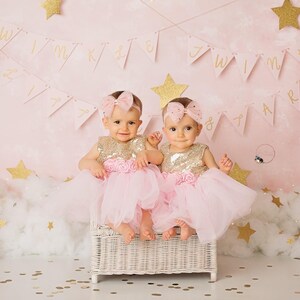 Op de afbeelding: Twee babymeisjes in roze tulejurken met gouden paillettentops en bijpassende roze strikken in hun haar. Ze zitten op een witte rieten stoel voor een roze achtergrond met gouden glittersterren en een banner met de tekst "Twinkle Twinkle Little Star".