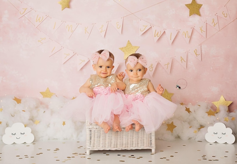 Op de afbeelding: Twee babymeisjes in roze tulejurken met gouden paillettentops en bijpassende roze strikken in hun haar. Ze zitten op een witte rieten stoel voor een roze achtergrond met gouden glittersterren en een banner met de tekst "Twinkle Twinkle Little Star".