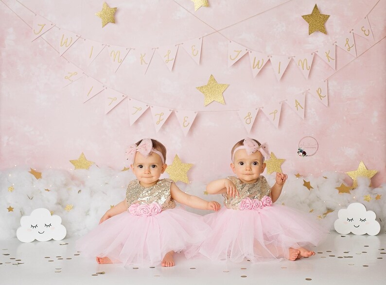 Op de afbeelding: Twee babymeisjes die roze tulejurken dragen met gouden pailletten-bovenstukken en roze bloemenaccenten. Ze zitten op een wit, pluche oppervlak met gouden glitterconfetti en een roze achtergrond met gouden glittersterren en een banner met de tekst "Twin Birthday".