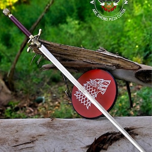 May include: A long sword with a dark red handle, a metallic crossguard, and a silver blade. Behind the sword is a circular red pizza cutter featuring a white wolf head emblem. The text AL MAJID PRODUCTION AXE, SWORD, PIZZA CUTTER is visible.