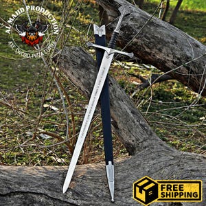 Puede incluir: Una espada plateada con una vaina negra, con un mango decorativo y una empuñadura en forma de estrella. La espada está apoyada en una rama de árbol con un fondo verde y marrón.