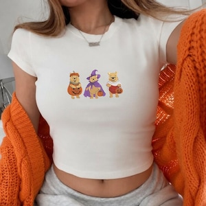 Puede incluir: Una camiseta blanca de manga corta con un estampado gráfico de tres osos amarillos con disfraces de Halloween. El primer oso está dentro de una calabaza, el segundo lleva un sombrero y una capa de bruja morados, y el tercero está cubierto con una sábana de fantasma blanca.