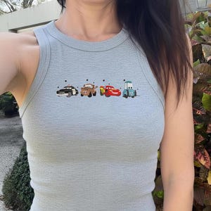 May include: A light gray ribbed tank top featuring a graphic print of four cartoon cars from the movie Cars, including a black police car, a brown tow truck, a red race car, and a light blue vehicle, arranged horizontally across the chest.