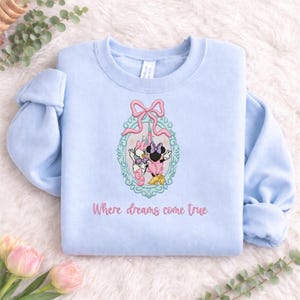 Puede incluir: Una sudadera azul claro de cuello redondo con un marco ovalado bordado con un lazo rosa en la parte superior. Dentro del marco, Daisy Duck y Minnie Mouse est&aacute;n frente a un castillo. El texto "Where dreams come true" est&aacute; bordado debajo.