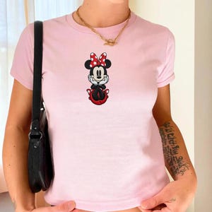 May include: A light pink t-shirt featuring a Minnie Mouse graphic. The cartoon character is depicted with a red and white polka dot bow, black ears, and a red skirt. The shirt has short sleeves and a crew neck.