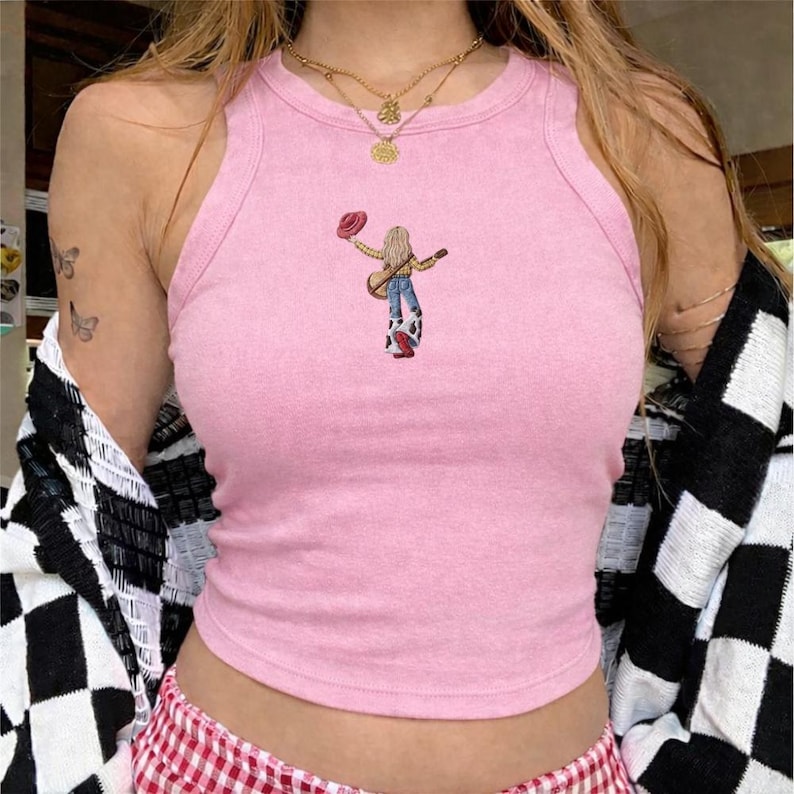May include: A light pink sleeveless tank top featuring a small embroidered graphic of a person with long blonde hair wearing a yellow shirt, blue jeans, and cow-print chaps, holding a red hat and a guitar, with a gold layered necklace.