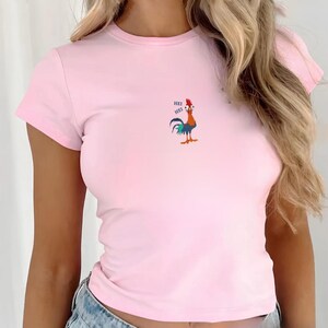 May include: A light pink t-shirt with short sleeves. The shirt features a colorful cartoon rooster with the text "HEI" above it. The rooster has a red comb, blue and green tail feathers, and is standing upright.