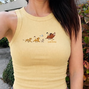 May include: A pale yellow ribbed tank top featuring a graphic print of characters from The Lion King, including Simba, Timon, Pumbaa, and Zazu, with the text Disney The Lion King printed in a small brown font below the animated figures.