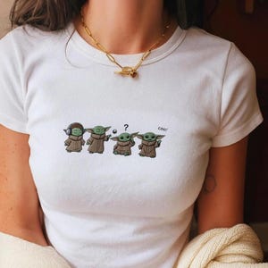 May include: White t-shirt featuring a cartoon design of four Grogu characters in different poses. The first Grogu wears headphones, the second holds a ball, the third has a question mark, and the last says "cool!"