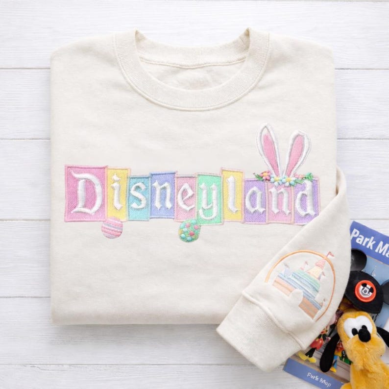 K&ouml;nnte beinhalten: Cremefarbenes Sweatshirt mit dem gestickten Wort "Disneyland" in Pastellfarben. Das Design umfasst Hasenohren und Ostereier. Der &Auml;rmel zeigt ein Schlossdesign. Eine Parkkarte und ein Pl&uuml;schtier sind sichtbar.