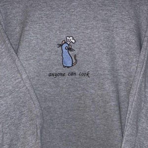 May include: A gray sweatshirt with a blue embroidered rat wearing a chef's hat. The text "anyone can cook" is embroidered below the rat. The sweatshirt has long sleeves and a crew neck.