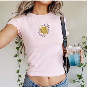 May include: A light pink t-shirt featuring a gold sun design with purple floral accents. The short-sleeved shirt is made of a soft material and is a casual top. The sun graphic is embroidered on the front.
