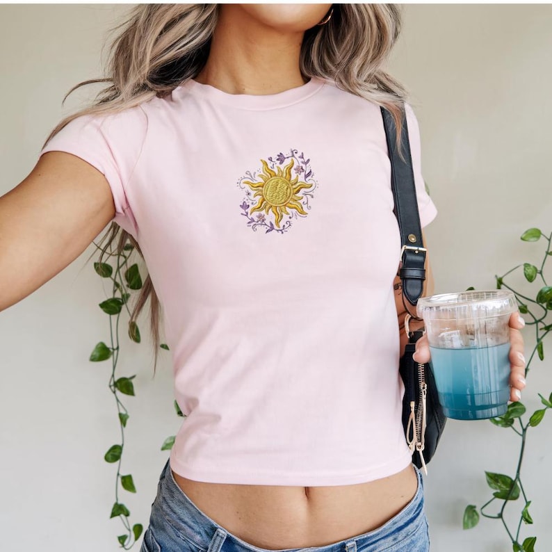 May include: A light pink t-shirt featuring a gold sun design with purple floral accents. The short-sleeved shirt is made of a soft material and is a casual top. The sun graphic is embroidered on the front.