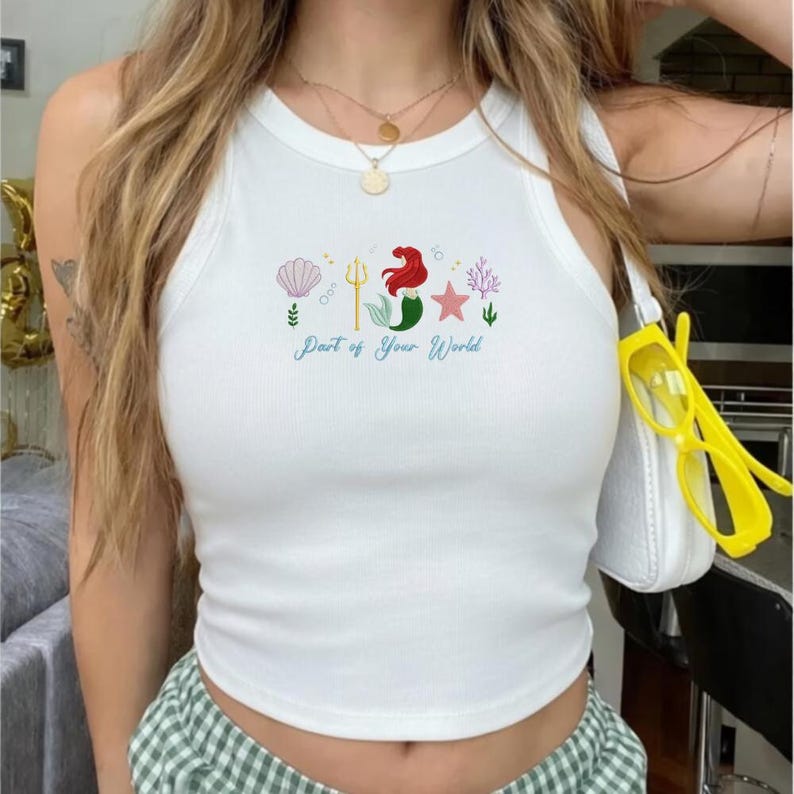 Puede incluir: Camiseta sin mangas blanca con un dise&ntilde;o bordado colorido que presenta una sirena, una concha, una estrella de mar y coral. El texto "Part of Your World" est&aacute; escrito en una fuente de guion debajo. La parte superior es sin mangas.