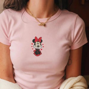 Peut inclure: T-shirt rose clair avec un motif de Minnie Mouse. Le dessin montre Minnie Mouse dans une robe et un n&oelig;ud rouges, entour&eacute;e de petits c&oelig;urs rouges. Le t-shirt a un col rond et des manches courtes.
