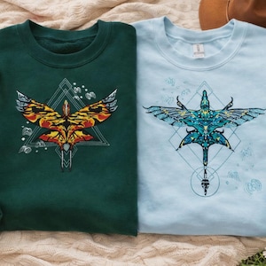 May include: Two sweatshirts, one dark green and one light blue, each featuring a detailed graphic of a stylized bird with geometric shapes. The green sweatshirt has a bird design in yellow, orange, and red. The blue sweatshirt has a blue and teal bird design.