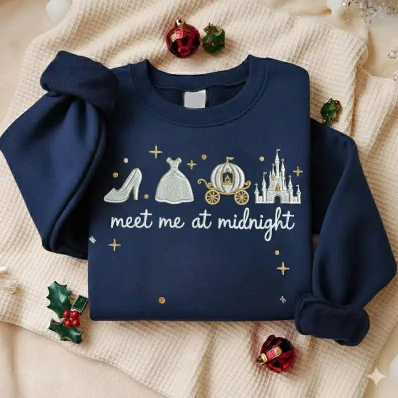 Puede incluir: Sudadera azul marino con un dise&ntilde;o tem&aacute;tico de Cenicienta. El dise&ntilde;o incluye un tac&oacute;n alto, un vestido de baile, un carruaje y un castillo, con el texto "meet me at midnight". La sudadera est&aacute; decorada con detalles dorados y cruces.