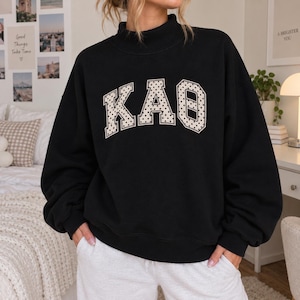 Könnte beinhalten: Ein schwarzes Langarm-Sweatshirt mit Stehkragen. Auf der Vorderseite befinden sich die griechischen Buchstaben KAO aus cremefarbenem Stoff mit schwarzem Punktemuster, die mit einem dicken cremefarbenen Rand versehen und mittig platziert sind.