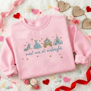 Puede incluir: Sudadera rosa claro con bordados de un zapato, un vestido, un carruaje y un castillo. El texto "meet me at midnight" est&aacute; bordado debajo. La sudadera est&aacute; rodeada de corazones y flores.