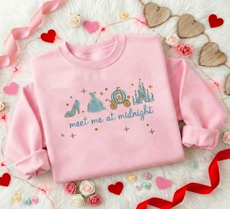 Puede incluir: Sudadera rosa claro con bordados de un zapato, un vestido, un carruaje y un castillo. El texto "meet me at midnight" est&aacute; bordado debajo. La sudadera est&aacute; rodeada de corazones y flores.